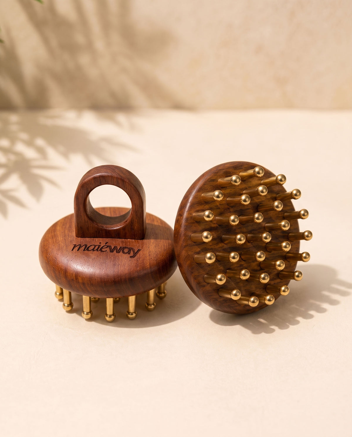 Two wooden Kansa scalp massagers with copper massage tips, one showing the handle side engraved with 'maiéway' and the other showing the brush head, set on a beige surface with soft shadows.