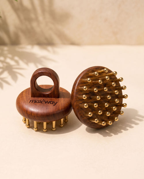 Two wooden Kansa scalp massagers with copper massage tips, one showing the handle side engraved with 'maiéway' and the other showing the brush head, set on a beige surface with soft shadows.