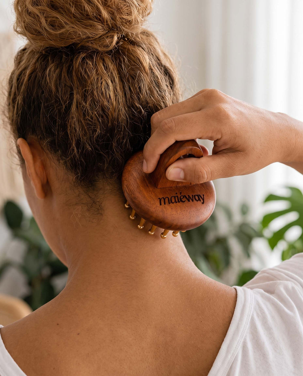 A woman seen from behind using a wooden scalp massager on the back of her neck. The product is branded with 'maiéway' in black lettering on the rose wood head, and small copper massage pins are visible along the bottom edge.