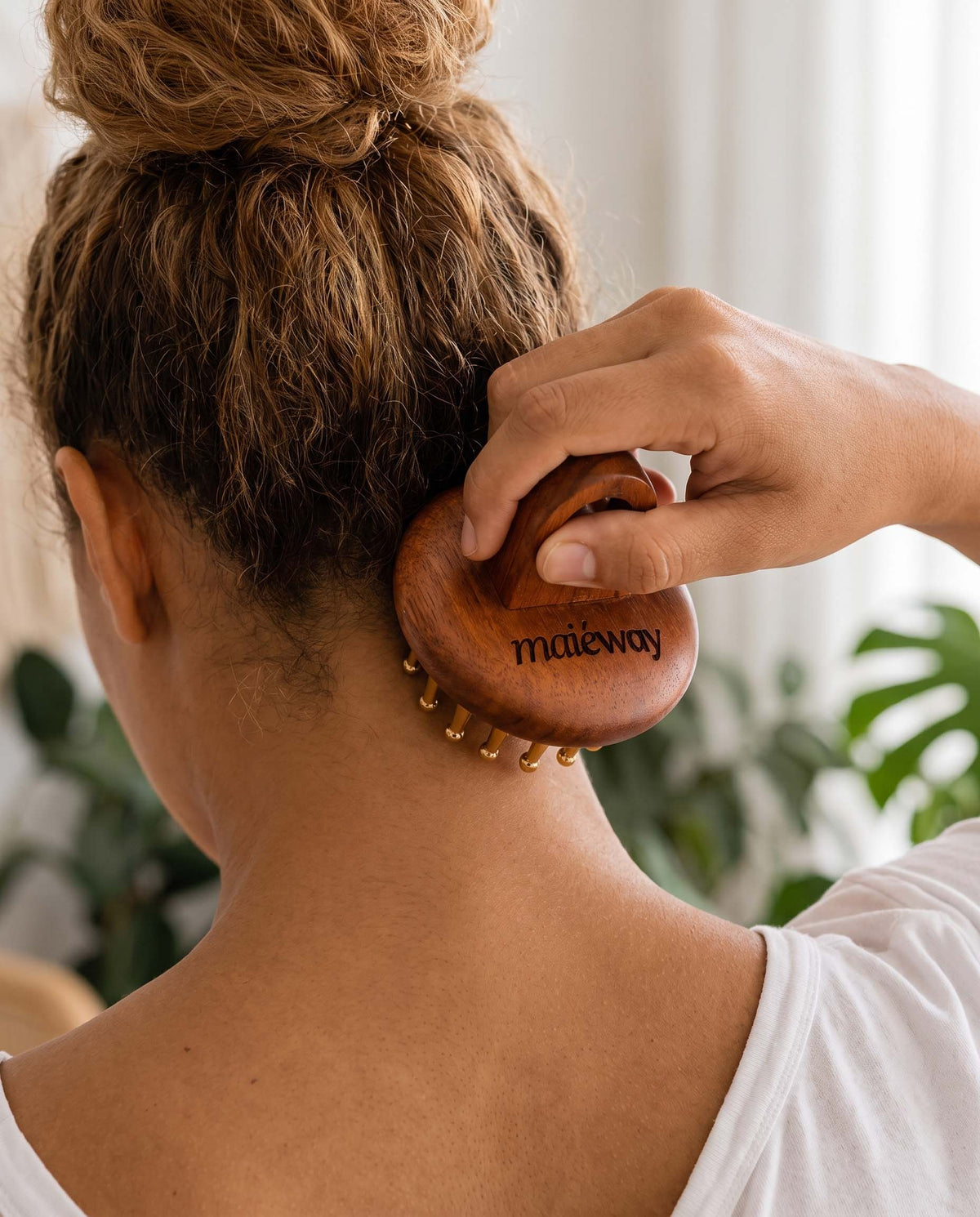A woman seen from behind using a wooden scalp massager on the back of her neck. The product is branded with 'maiéway' in black lettering on the rose wood head, and small copper massage pins are visible along the bottom edge.
