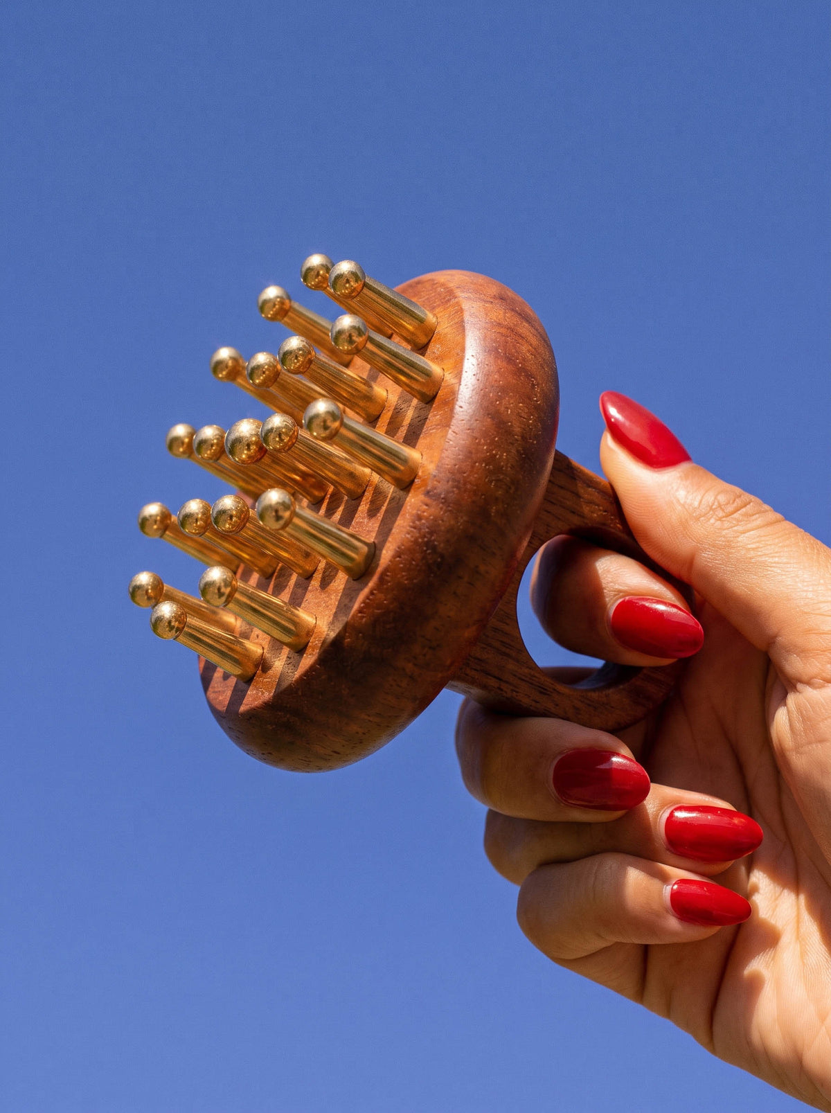 A hand with red painted nails holds a Kansa Rosewood scalp massager with copper massage pins against a clear blue sky background.