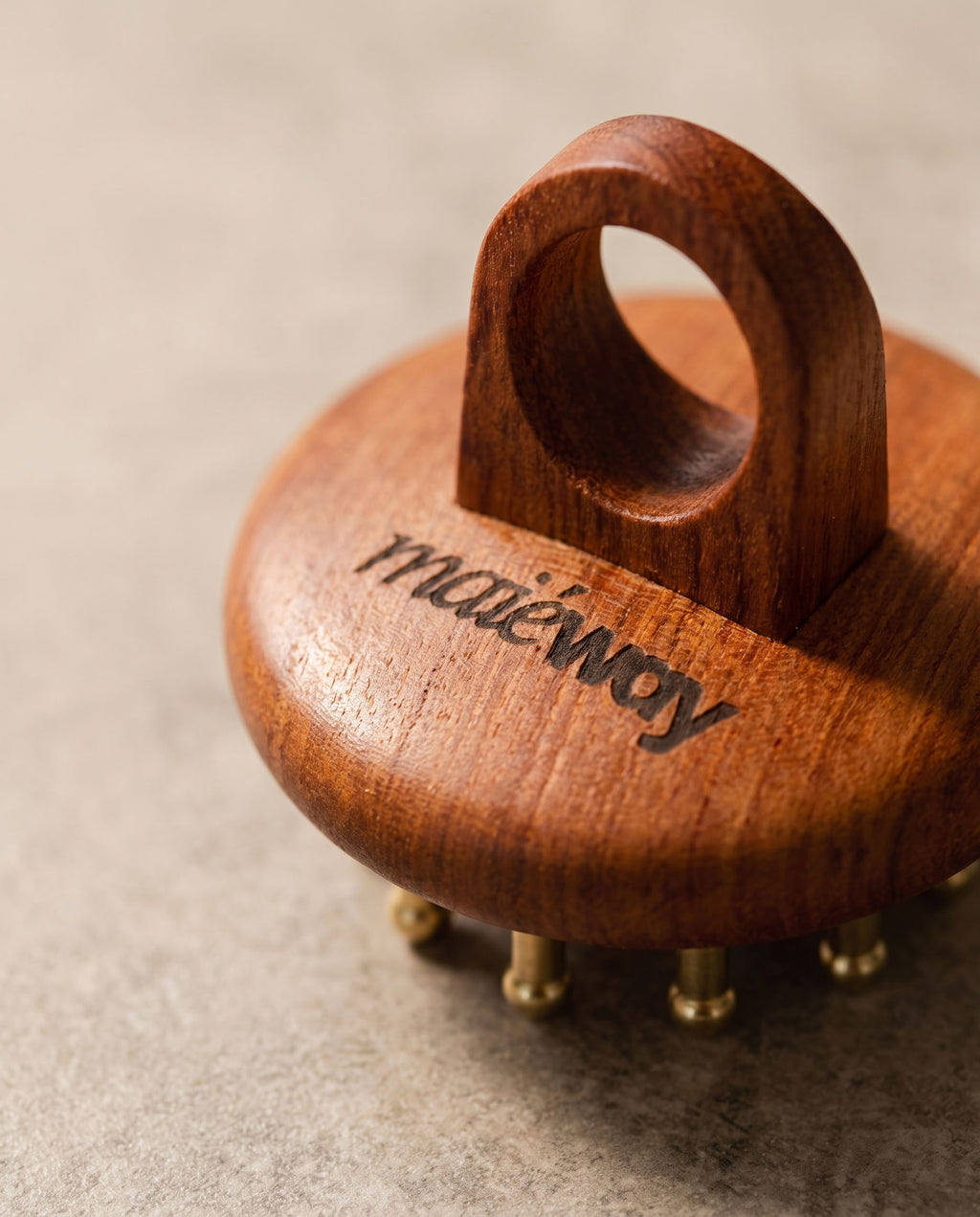 A close-up of a Kansa scalp massager with a loop handle and small copper prongs, featuring the 'maiéway' brand name engraved on the side, set against a neutral textured background.