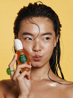 A close-up lifestyle shot of a model with wet hair holding a bottle of Scalp Prime 5 Scalp Primer against a solid yellow background.