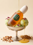 A bottle of Maieway Scalp Primer filled with golden hair oil, propped up in a silver pedestal bowl with green ice cream scoops,  Scalp Primer oil dripping over the scene, almonds, and fenugreek on a warm beige background.